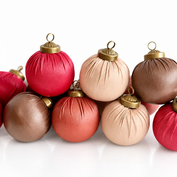 A group of decorative Christmas ornaments shaped like round balls, shown on a clean white background. The ornaments come in red, coral, blush pink, and taupe-brown satin-like finishes, each with a gold-tone cap and hanging loop.