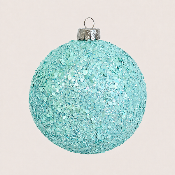 A round Christmas ornament covered in light blue glitter, featuring a silver metal cap and hanging loop, set against a solid pale pink background.