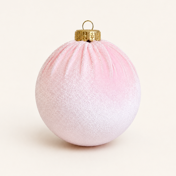 A spherical Christmas ornament with a textured fabric-like surface, featuring a soft pink to white ombré gradient and a metallic gold cap with a hanging loop, set against a plain off-white background.