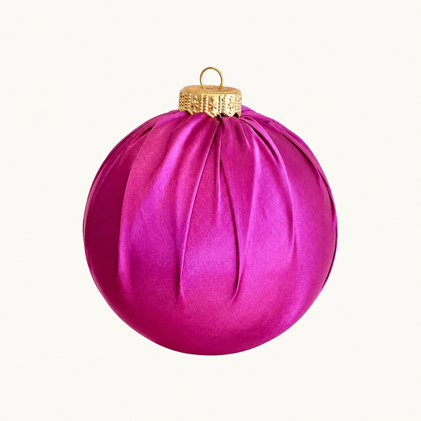 A spherical Christmas ornament in a vibrant magenta pink satin fabric, featuring a textured gold cap and a small gold hanging loop, set against a plain off-white background.