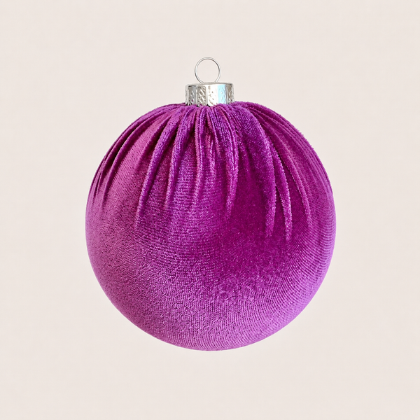 A spherical Christmas ornament in textured purple velvet with a silver metal cap and hanging loop, centered against a solid light beige background.