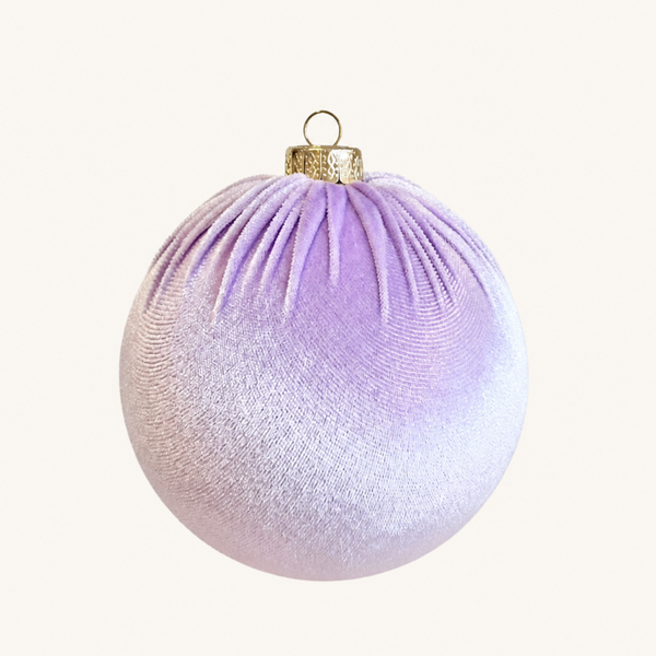 A spherical Christmas ornament with a textured, ribbed fabric-like surface, featuring a lavender purple to pale pink gradient and a gold hanging cap at the top, shown against a solid off-white background.
