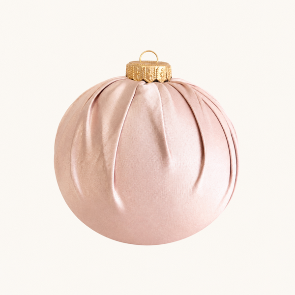 A ribbed, pleated Christmas ornament in a soft blush pink satin-like finish with a textured gold cap and hanging loop, shown against a solid off-white background.