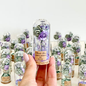 Hand holding a small glass bottle with preserved flowers and a personalized label against a background of similar bottles.