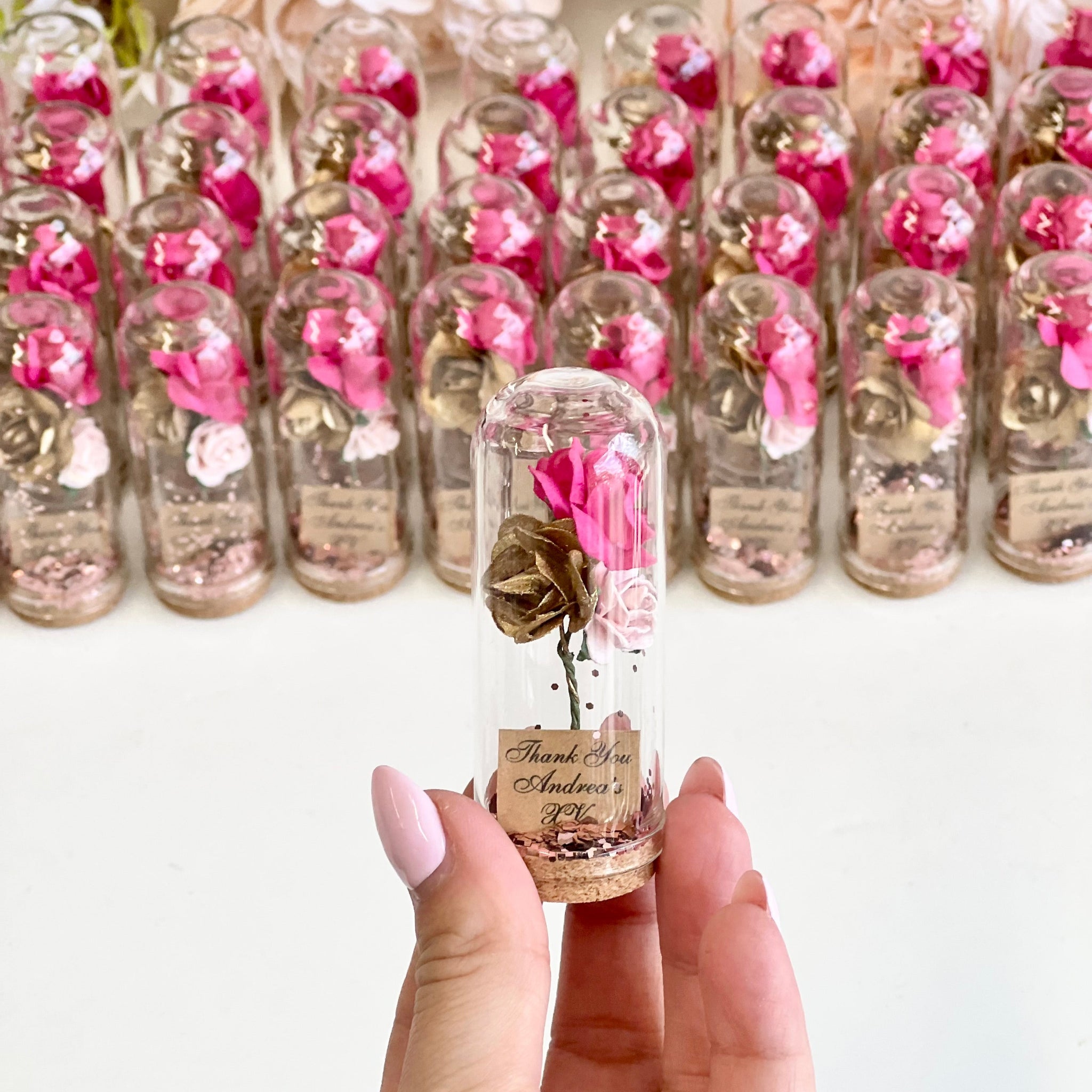 Decorative vials with pink flowers and gold ribbons held by a hand, with more vials in the background.