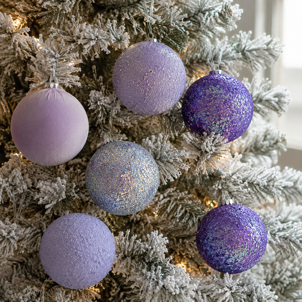 Six decorative Christmas tree ornaments in shades of lavender, purple, and glittery blue, hanging on a flocked white Christmas tree with warm string lights.