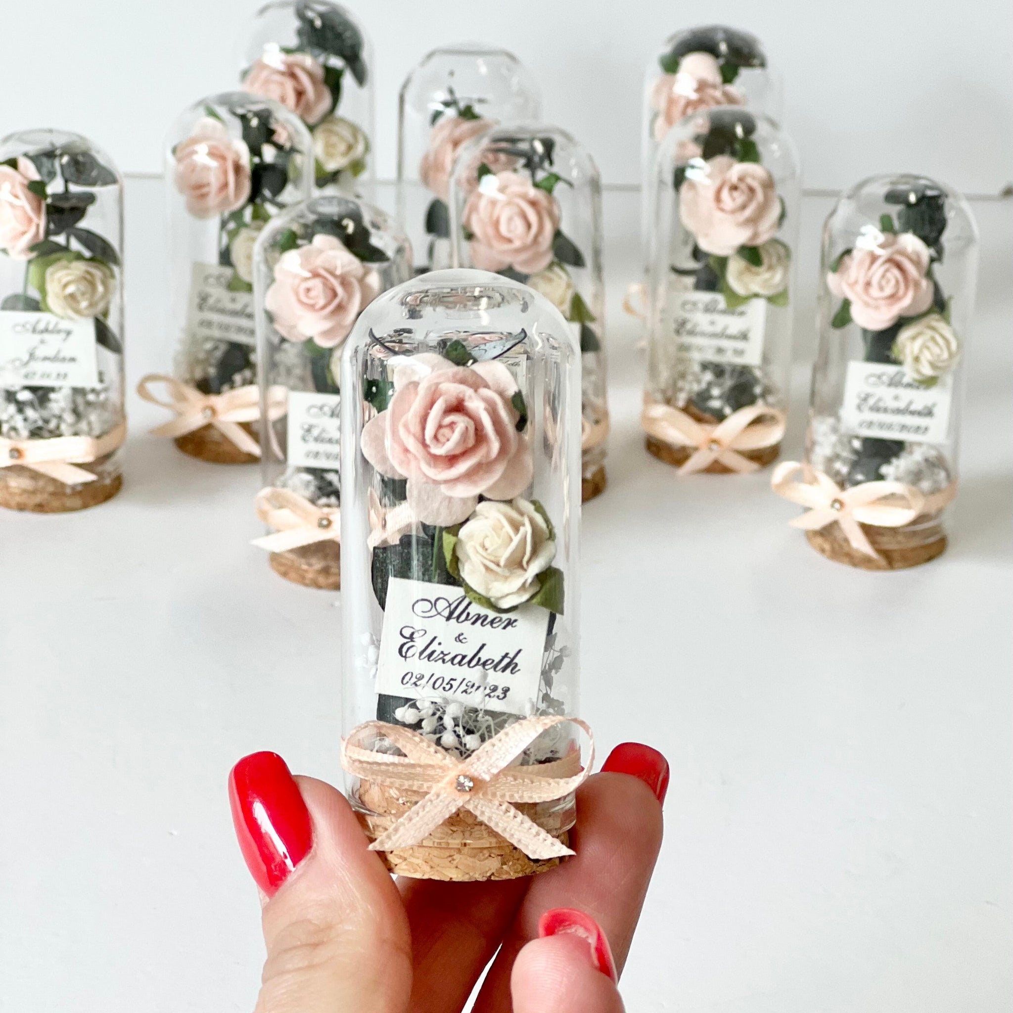Small glass bottles with preserved flowers and labels held by a hand against a white background