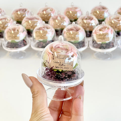 Hand holding a small cake in a clear dome with more cakes in the background.
