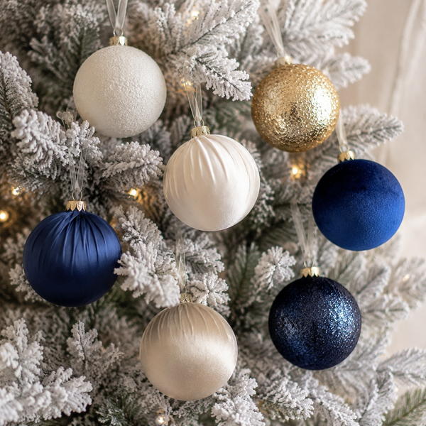 A set of seven Christmas tree ornaments in white, gold, and navy blue, hanging on a flocked white Christmas tree with warm string lights.
