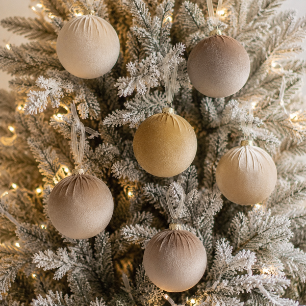 Six matte fabric Christmas tree ornaments in neutral beige and gold shades, hanging on a frosted white Christmas tree with warm string lights.
