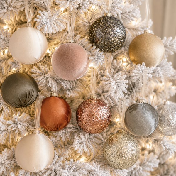 A close-up of white-flocked Christmas tree branches decorated with multiple matte and glitter holiday ornaments in neutral and metallic tones, including cream, blush, taupe, rose gold, copper, silver, and gold.