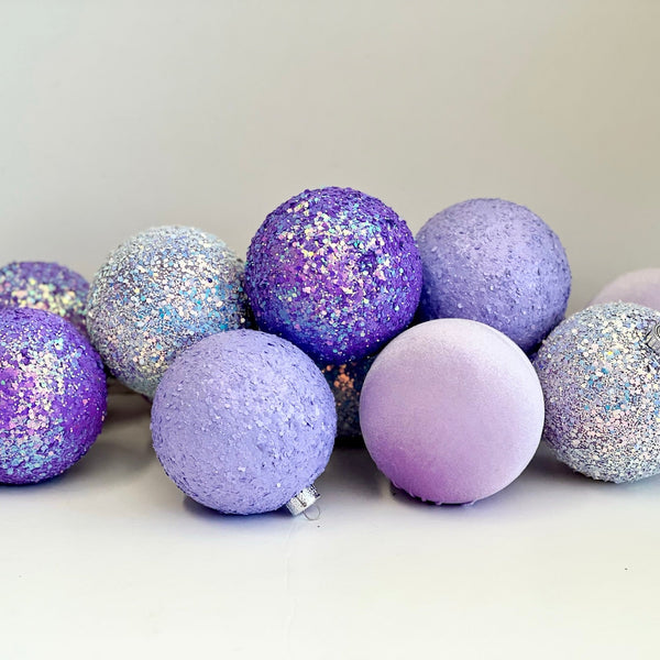 A group of decorative spherical ornaments in pastel purple, lavender, and glittery silver, arranged on a plain light background.