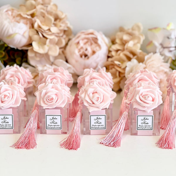 A row of small pink flower boxes with pale pink roses, pink tassels, and white labels, arranged in front of large beige and cream flowers against a light neutral background.