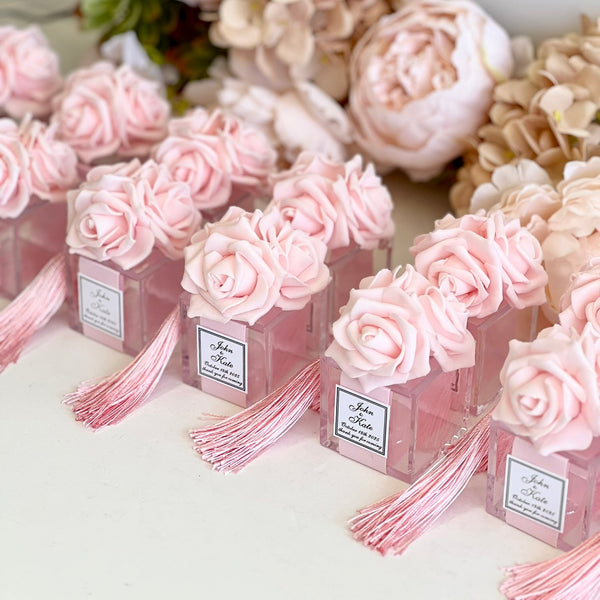 Pink rose-shaped perfume bottles with pink tassels and white labels, arranged on a white surface with dried flowers in the background.