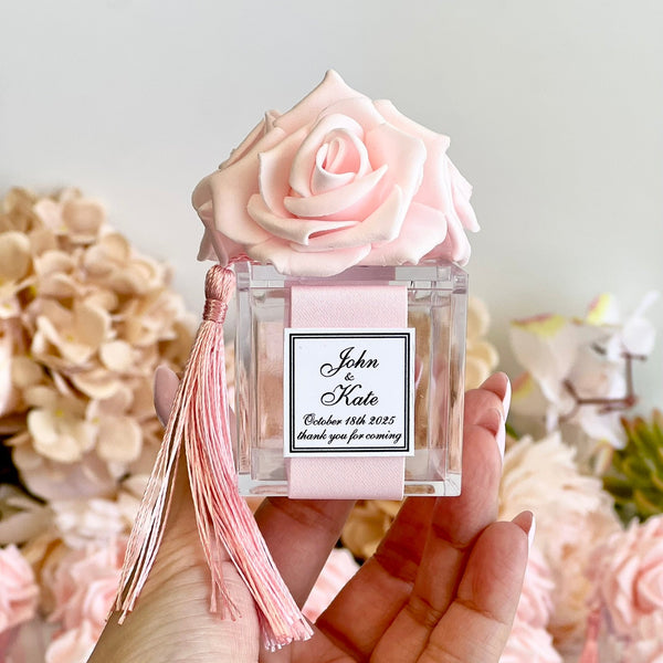 A hand holds a clear glass perfume bottle decorated with a large light pink rose on top, a matching pink ribbon and tassel, and a custom printed white label. The background is softly blurred with more pink roses and floral arrangements.