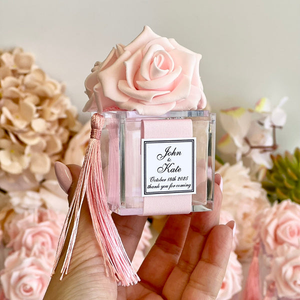 A hand holds a personalized pink rose diffuser bottle with a pink tassel. The clear glass bottle features a light pink artificial rose on top and a custom white label that reads 'John & Kate' with a wedding date.