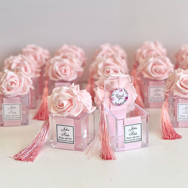A row of pink rose-shaped favor boxes with pink tassels and clear acrylic bases, each labeled with a personalized name and date, arranged on a light surface against a soft light gray background.