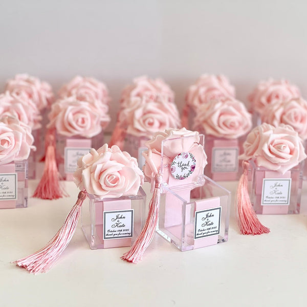 A set of pink rose favor boxes with pink tassels and clear acrylic bases, arranged on a light surface against a soft light gray background.