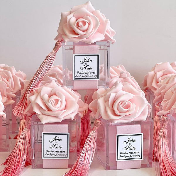 A stack of clear acrylic favor boxes decorated with large light pink roses and long pink tassels, each with a personalized wedding favor label reading 'John & Kate' and 'Thank you for coming.'.