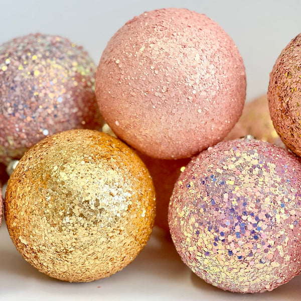 A close-up of several glittery decorative spheres in pastel pink, peach, gold, and lavender tones on a light neutral background.
