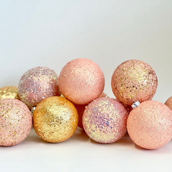 A group of glittery Christmas ornaments in pastel pink, peach, and metallic gold, arranged on a plain light background.