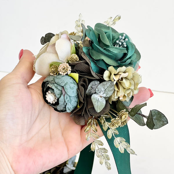 Hand holding a bouquet of artificial flowers with green and beige tones on a white background