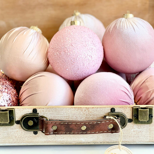 A coordinated set of twelve handmade Christmas ornaments in soft shades of pink, blush, and rose gold. The ornaments feature a variety of textures, including glitter, satin, and velvet, arranged in a grid pattern against a clean white background. This romantic and feminine color palette adds a delicate, modern charm to holiday decor.