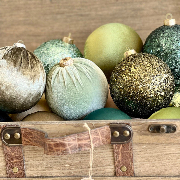 A vibrant 12-piece set of handmade Christmas ornaments in a gradient of green tones, from mint and olive to emerald and teal. The ornaments are made from a mix of glitter, satin, and velvet materials, arranged neatly in three rows by texture and color. This elegant and earthy color palette brings a modern and refreshing look to holiday decor.