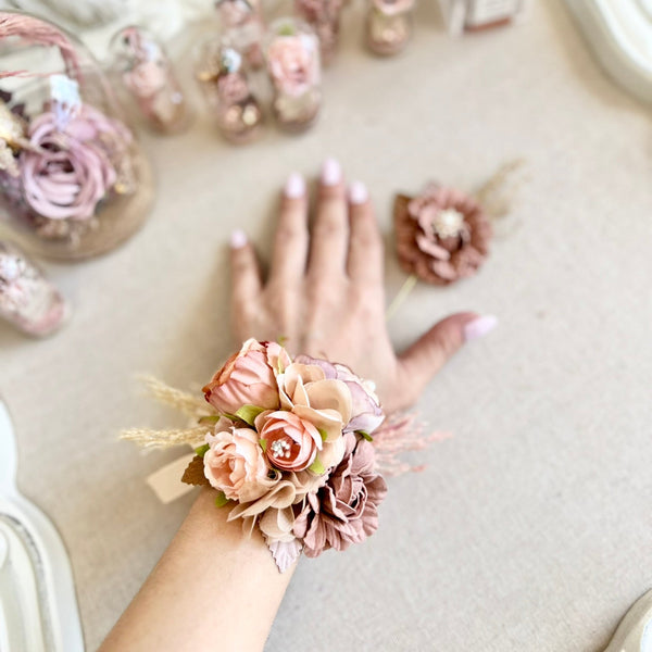 Hand wearing a floral wrist corsage with more corsages and flowers in the background.