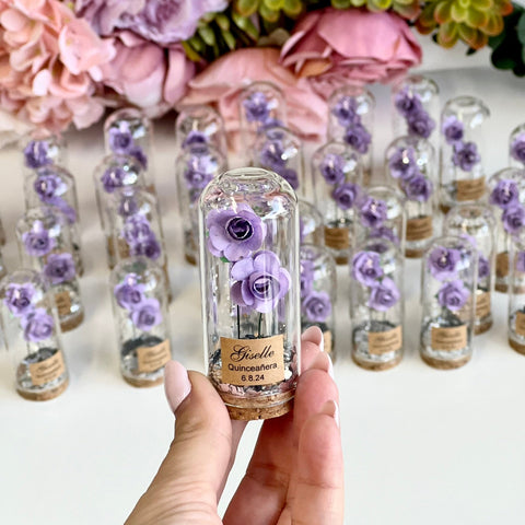 Hand holding a small glass bottle with preserved purple flowers, surrounded by more bottles and floral arrangements.