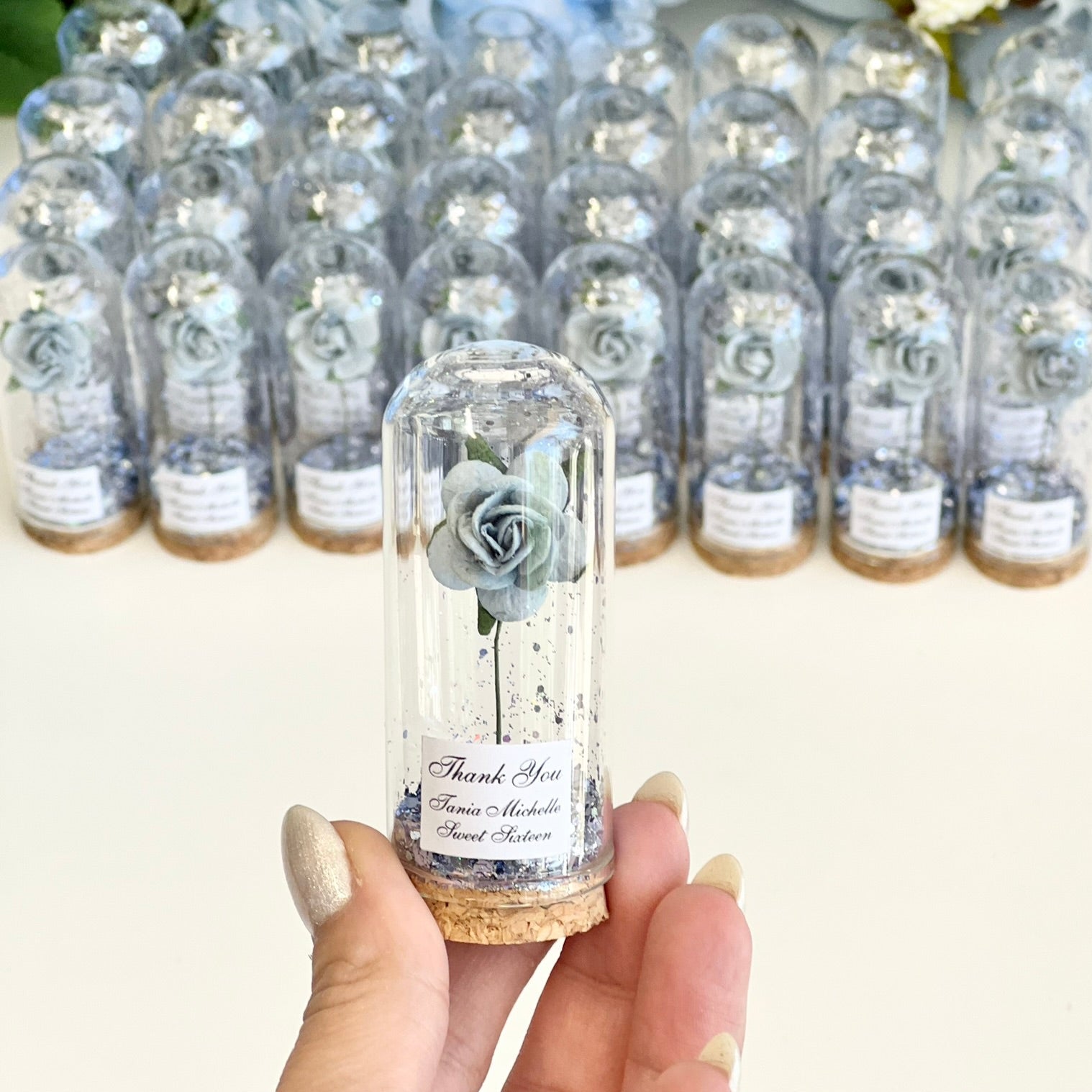 Clear glass bottles with small flowers inside, held by a hand against a floral background.
