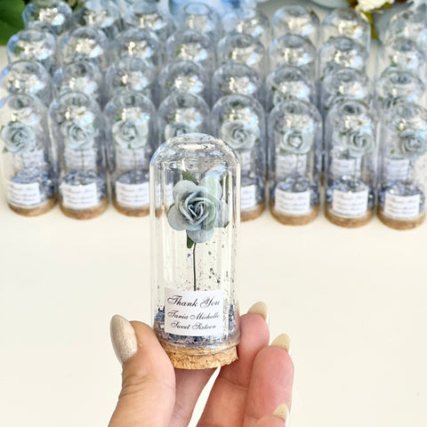Clear glass bottles with small flowers inside, held by a hand against a floral background.