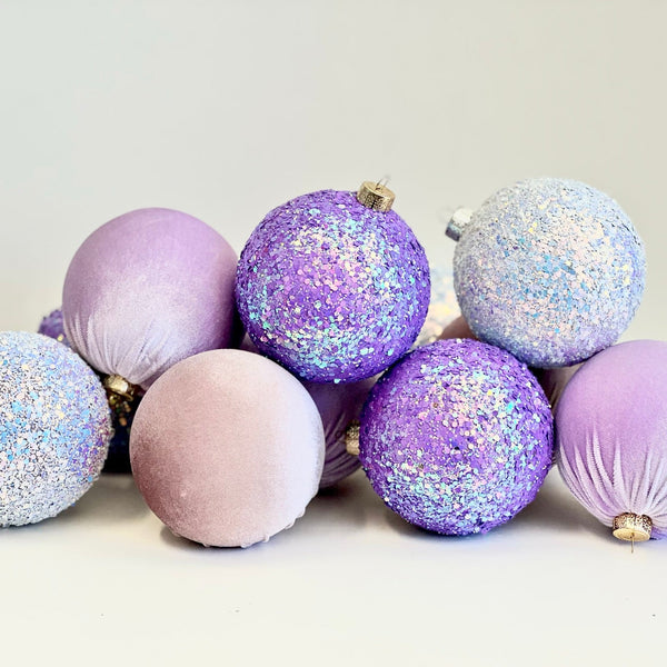 A group of decorative Christmas baubles in pastel lavender, light pink, and iridescent silver glitter, arranged on a plain light background.