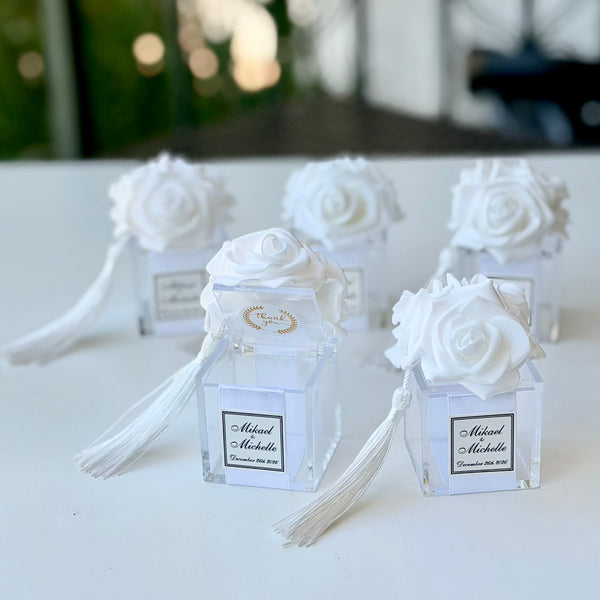 Five small clear glass favor boxes tied with white ribbons and topped with white fabric roses, each with a white tassel and a personalized label, arranged on a white table with a blurred outdoor background.