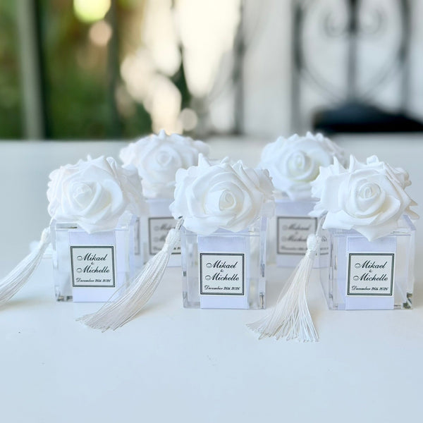 Five white rose favor boxes with white tassels and personalized labels, arranged on a white table with a blurred outdoor background.