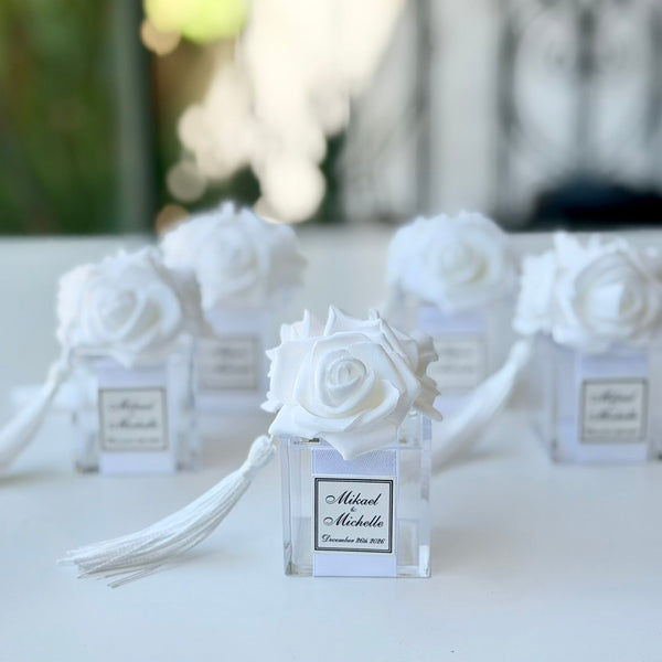 White fabric rose favor holders in clear square glass vials with white tassels, displayed on a white table with a softly blurred background.