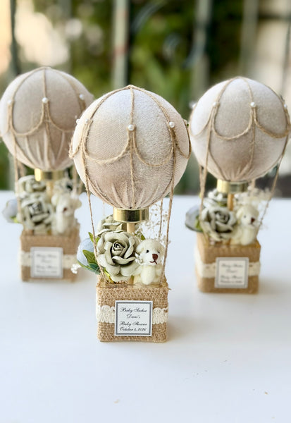 Three hot air balloon-themed baby shower favors arranged on a white surface. Each favor features a beige fabric balloon, burlap basket, faux roses, a small plush bunny, and a personalized name label.
