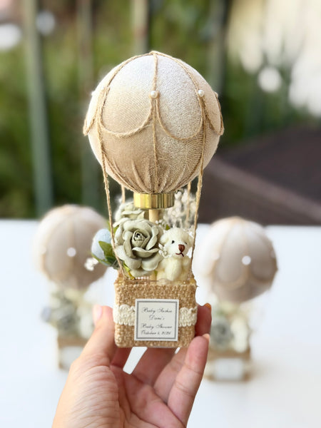A hand holds a decorative hot air balloon ornament with a beige fabric balloon, burlap-wrapped basket, faux flowers, and a small teddy bear. Two similar ornaments are blurred in the background on a white surface with an outdoor bokeh backdrop.