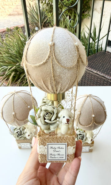 A hand holds a decorative hot air balloon ornament made of beige burlap fabric, twine, faux flowers, and a small teddy bear figure. Two matching balloon ornaments sit on a white table in the background, with a patio and plants visible behind them.