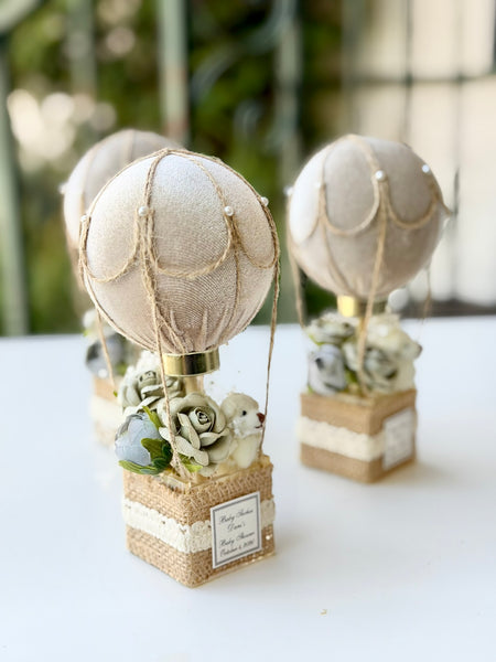 Three rustic hot air balloon table centerpieces with beige fabric balloons, burlap bases, faux flowers, and small white birds on a white tabletop.