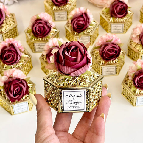 Decorative box with a rose and personalized label held by a hand, surrounded by similar boxes.