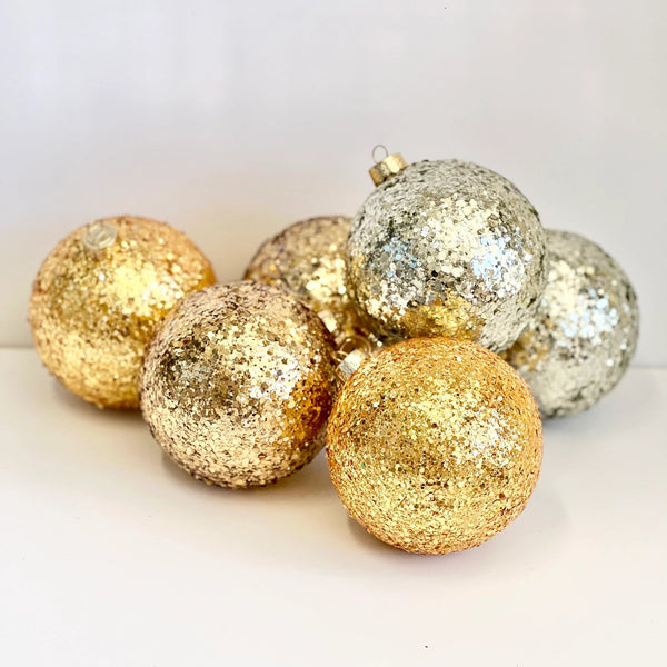 A group of glittery Christmas tree ornaments in metallic gold and silver, arranged on a plain white surface.
