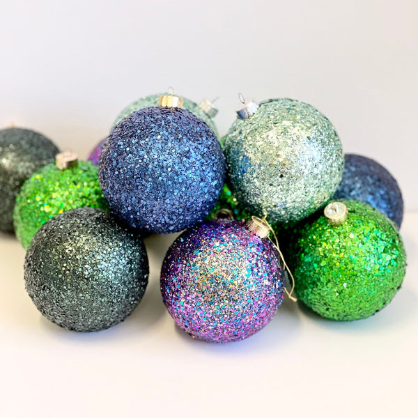 A pile of glitter Christmas ornaments in assorted colors including blue, green, purple, and silver, photographed on a clean white background.