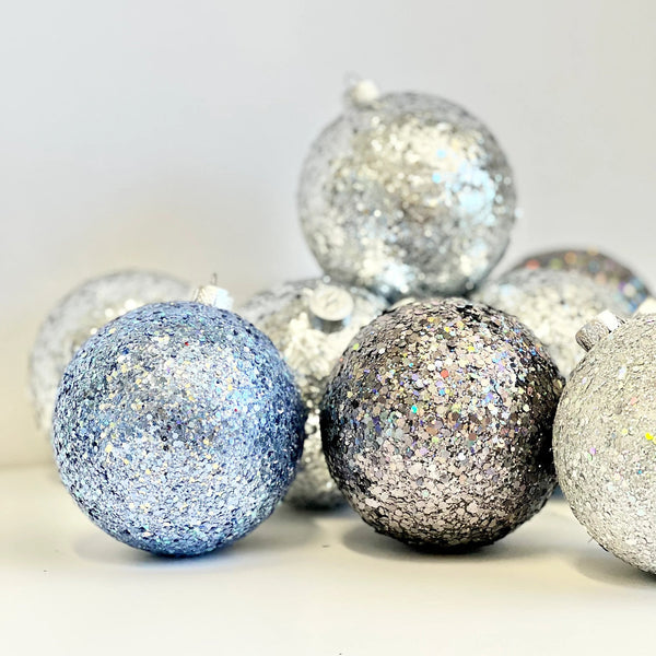A group of glittery Christmas ornaments in silver, white, and light blue, arranged on a plain light background.