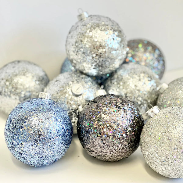 A pile of glittery Christmas ornaments in silver, light blue, and dark gray with iridescent sparkle, photographed on a plain light background.
