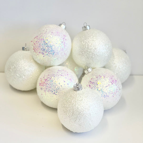 A group of white Christmas ornaments with textured glitter finishes, including some decorated with iridescent holographic glitter, arranged on a plain white surface.