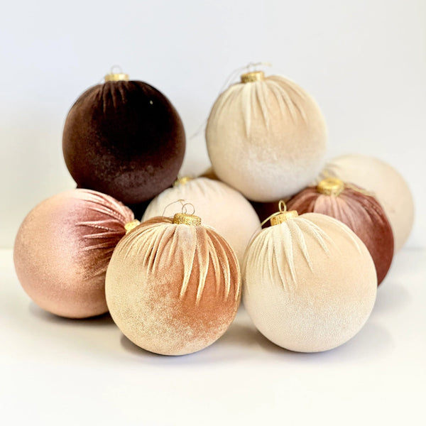 A group of velvet Christmas tree ornaments in muted neutral and warm metallic tones, including cream, blush pink, taupe, and deep brown, arranged on a white surface against a white background.