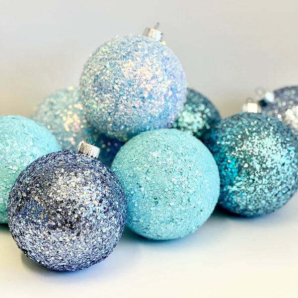 A group of glittery Christmas ornaments in shades of light blue, turquoise, and navy, arranged on a clean white surface against a light gray background.