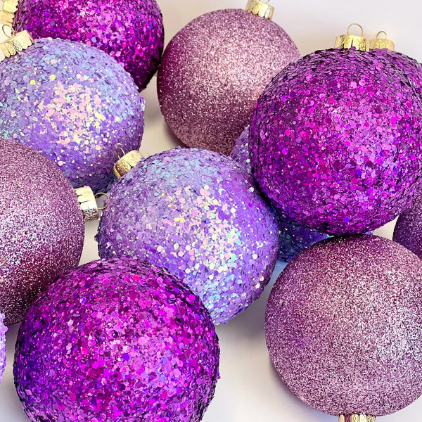 A group of glittery and textured purple Christmas ornaments with gold hanging loops, arranged closely together on a plain white background.