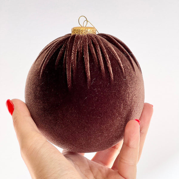 A hand with red painted nails holds a dark brown velvet Christmas bauble ornament with a gold hanging loop against a plain white background.